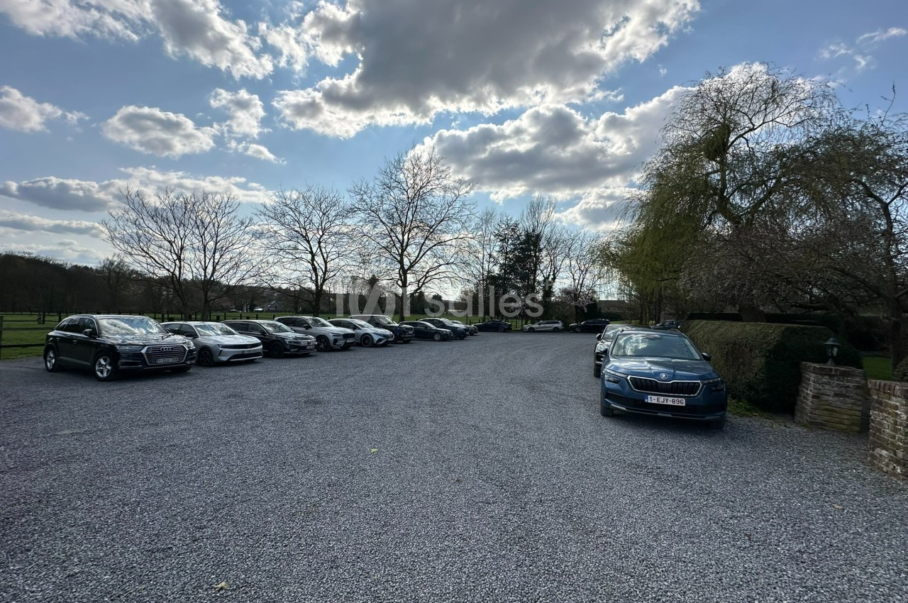 Parking extérieur avec plusieurs voitures stationnées sur un sol en gravier, entouré d'arbres et sous un ciel partiellement…