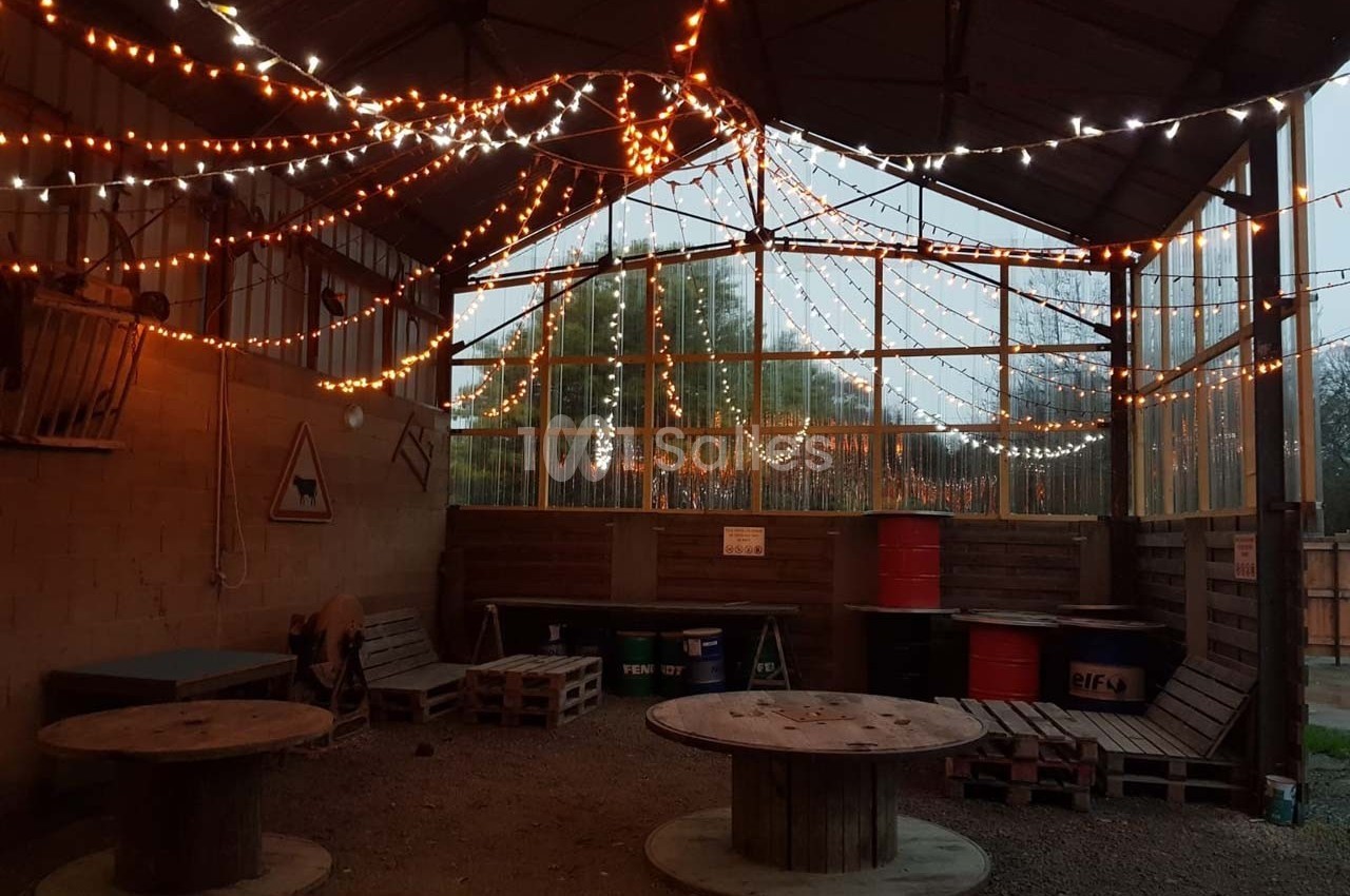 Espace couvert avec guirlandes lumineuses, tables en bois et palettes, ambiance rustique et chaleureuse.