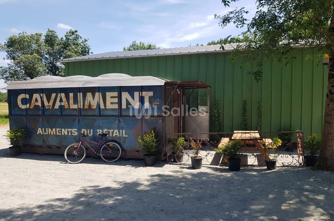 Un ancien wagon bleu transformé en point de vente, entouré de plantes en pot, avec une table en bois et un vélo rose devant.