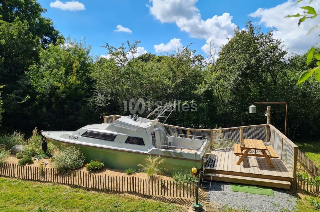 Un bateau aménagé en espace de détente est installé sur un terrain entouré de verdure avec une terrasse en bois.