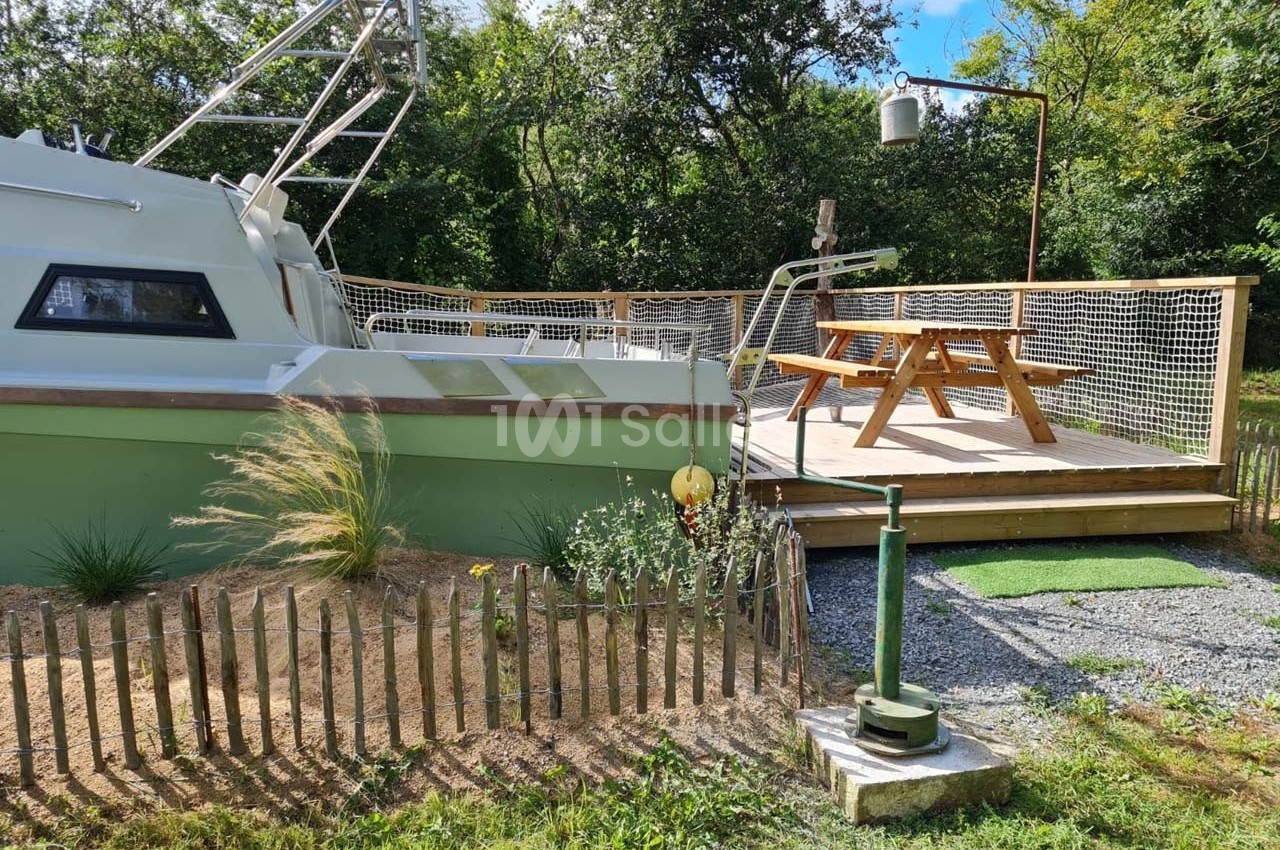 Bateau aménagé en hébergement avec terrasse en bois, table de pique-nique et environnement verdoyant.