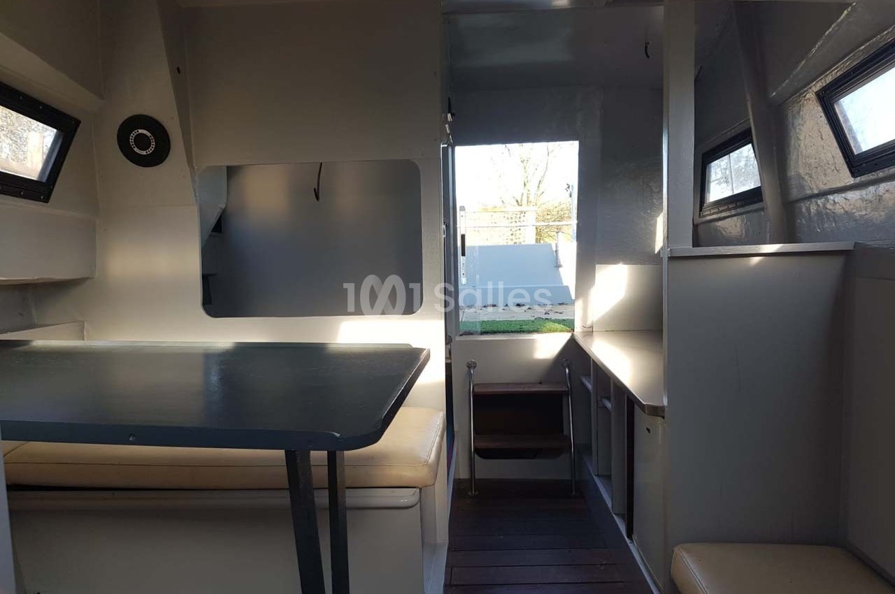 Intérieur d'un petit espace de vie avec table, banquettes, fenêtres et vue sur l'extérieur par une porte ouverte.