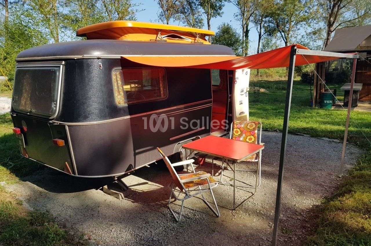Caravane vintage noire avec auvent orange, table et chaise pliantes sur un sol gravillonné, entourée de verdure.