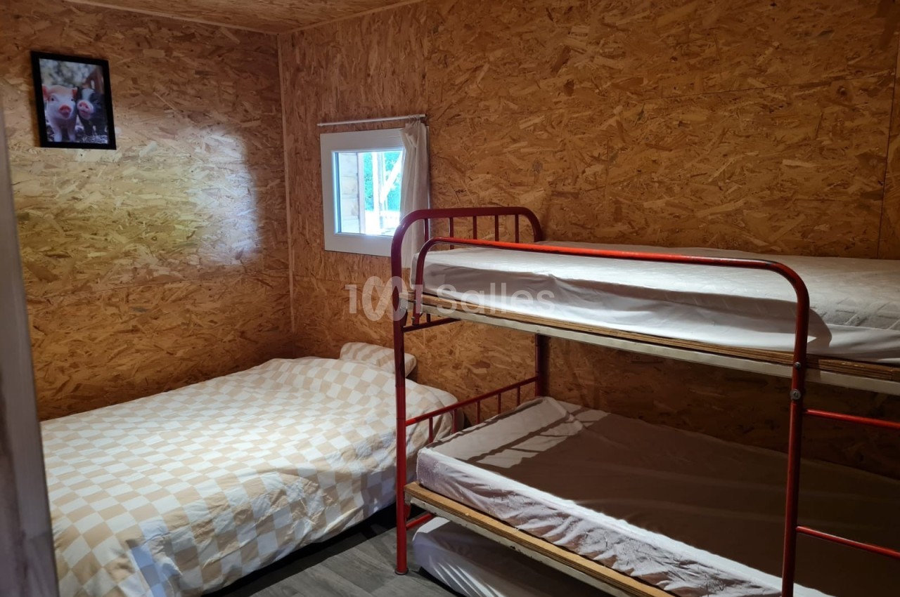 Chambre simple avec murs en bois, un lit simple, un lit superposé rouge et une petite fenêtre laissant entrer la lumière.