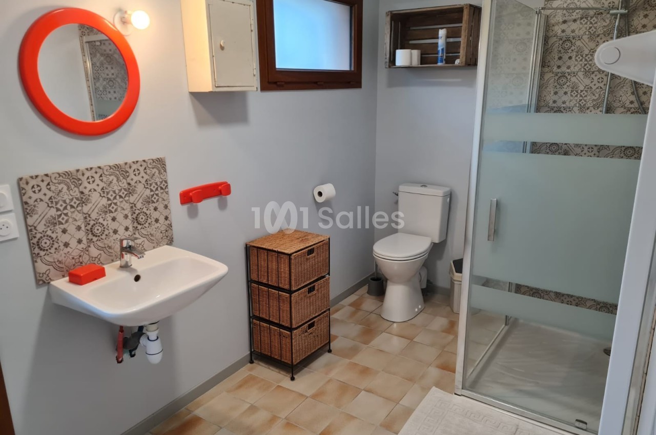 Salle de bain avec lavabo, miroir rond rouge, douche vitrée, toilettes et meubles en osier sur carrelage beige.