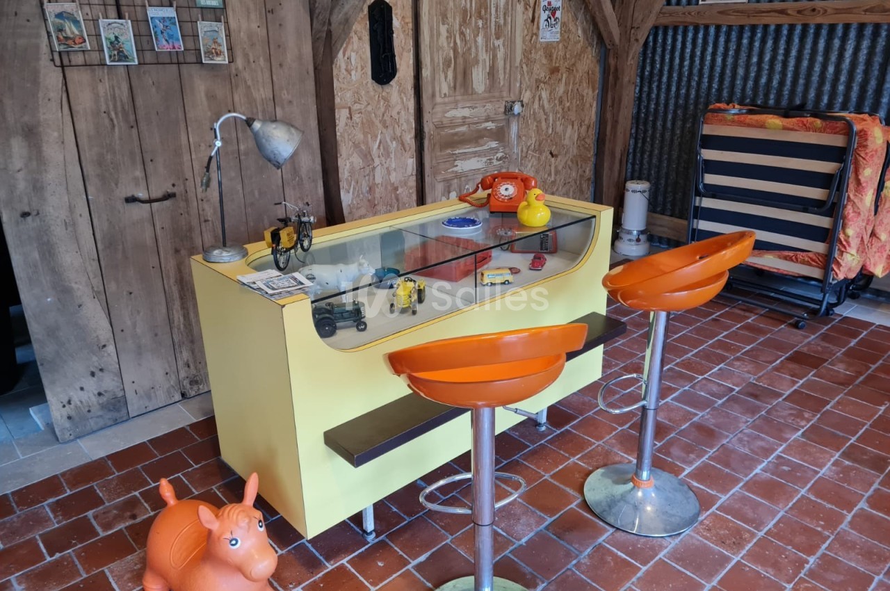 Comptoir jaune avec objets rétro, deux tabourets orange et un jouet en forme d'animal sur un sol en carrelage rouge.