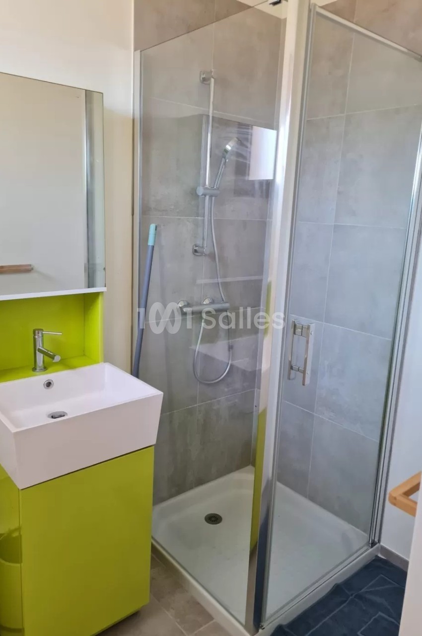 Salle de bain moderne avec douche vitrée, lavabo sur meuble vert et miroir, sol en carrelage clair.