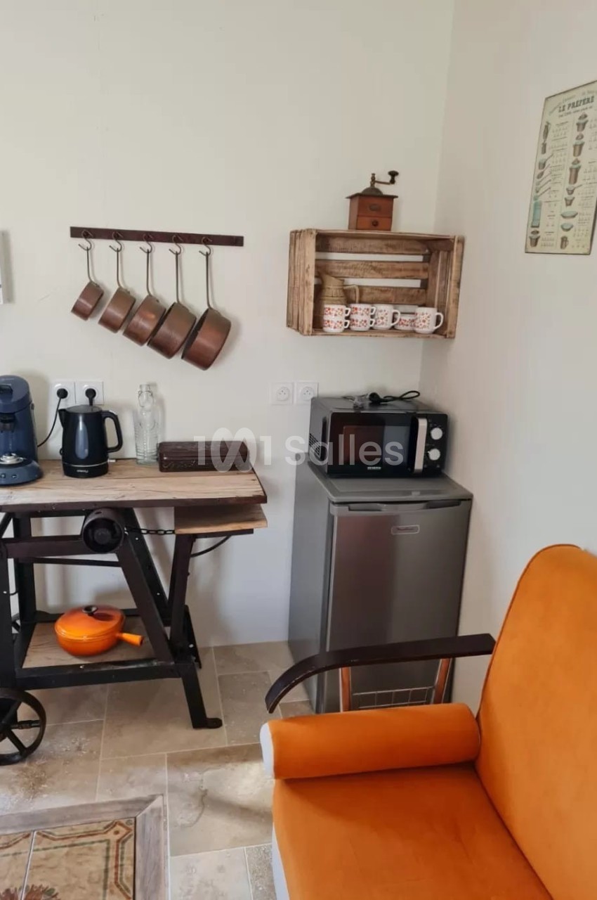 Coin cuisine avec cafetière, bouilloire, micro-ondes, casseroles suspendues, étagère en bois et fauteuil orange au premier…