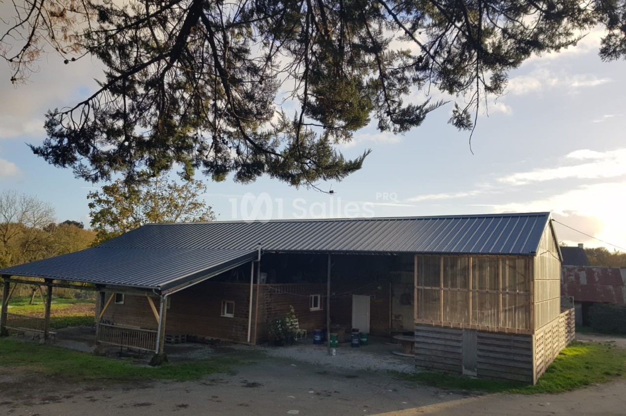 Bâtiment agricole en bois et métal avec un toit en pente, entouré d'arbres et baigné par une lumière naturelle.