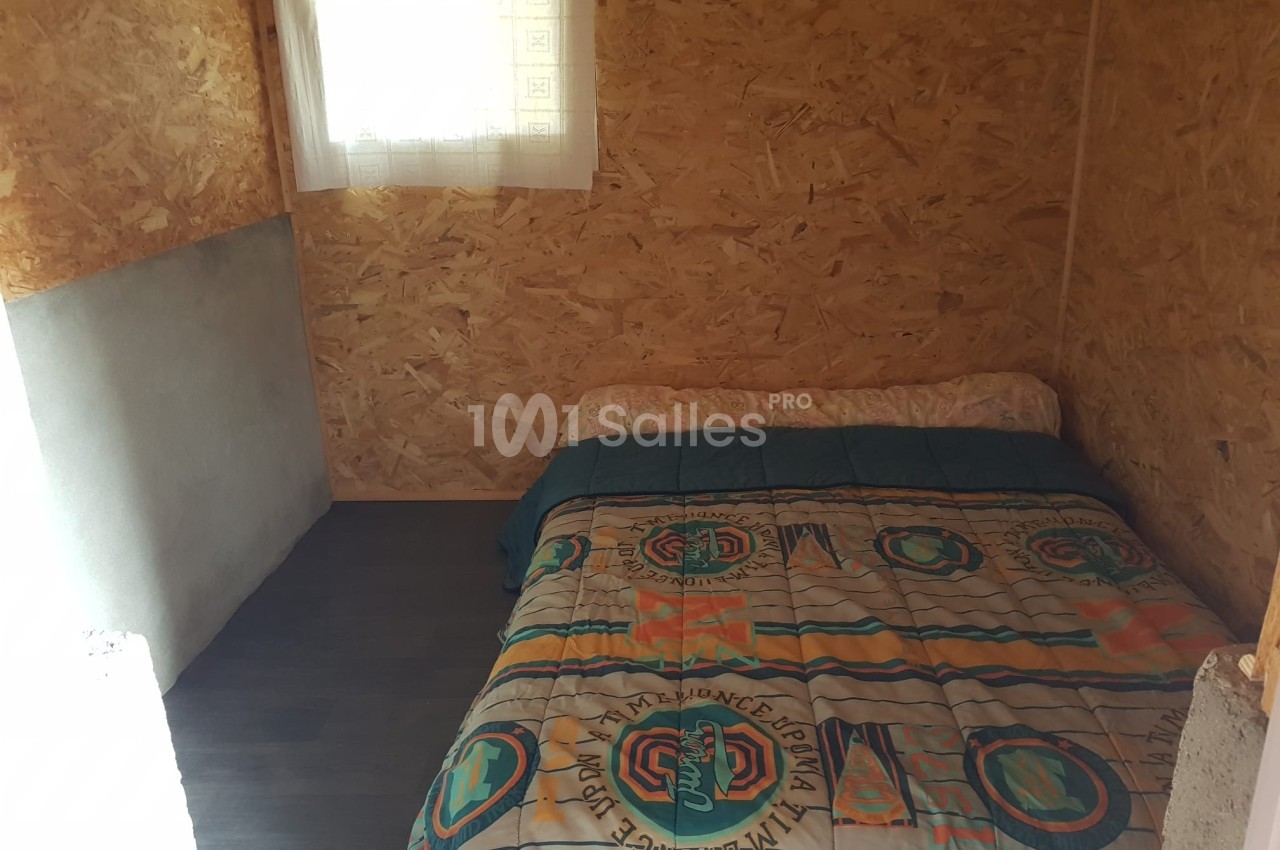 Lit double avec une couverture colorée dans une petite chambre aux murs en panneaux de bois, éclairée par une fenêtre.