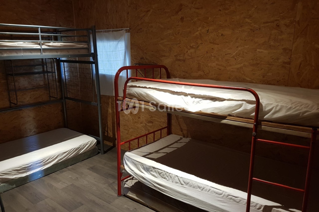 Chambre simple avec lits superposés en métal, murs en bois et fenêtre avec rideau blanc.