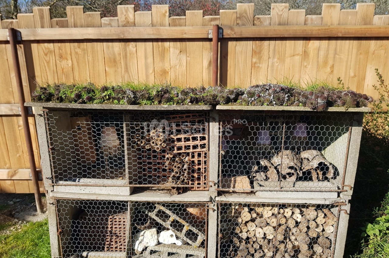 Hôtel à insectes en bois et grillage, rempli de matériaux naturels, installé dans un jardin près d'une clôture en bois.