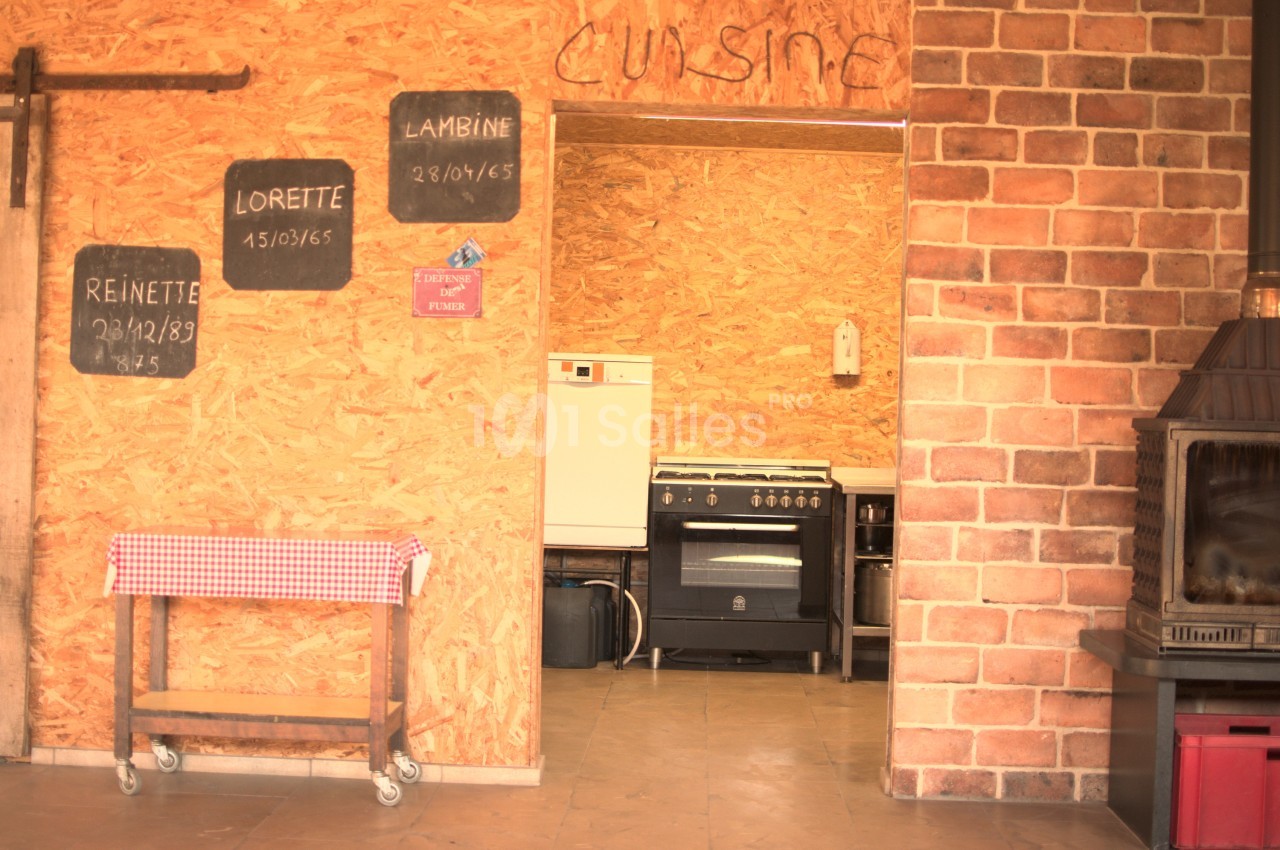 Cuisine rustique avec murs en bois et briques, table à nappe rouge, four et plaques de cuisson visibles en arrière-plan.