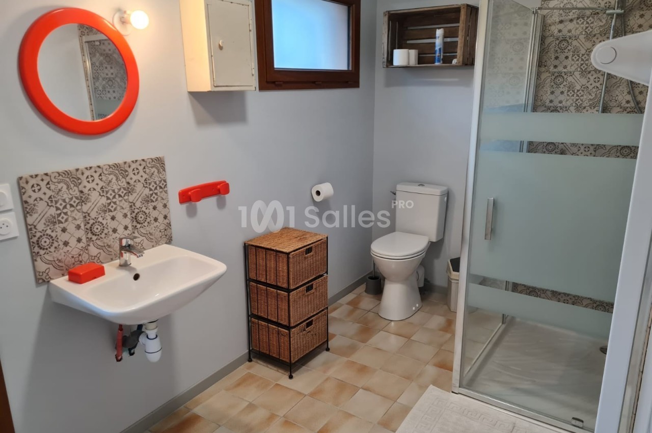 Salle de bain avec lavabo, miroir rond rouge, douche vitrée, toilettes et meubles de rangement en osier.