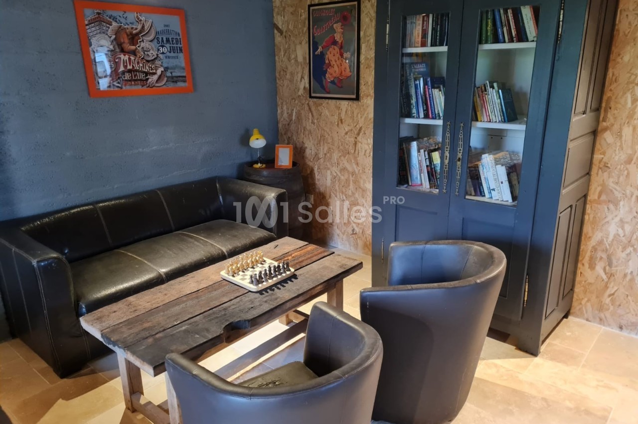 Espace de détente avec canapé, fauteuils, table basse avec échiquier, bibliothèque et décorations murales.