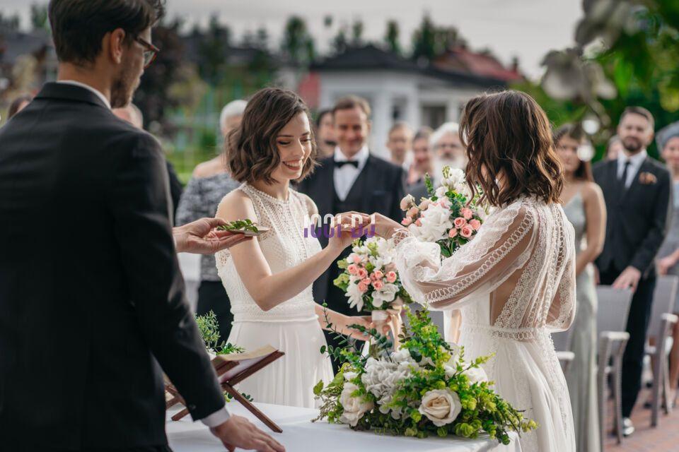 Deux femmes en robes blanches échangent des alliances lors d'une cérémonie de mariage en extérieur, entourées d'invités.