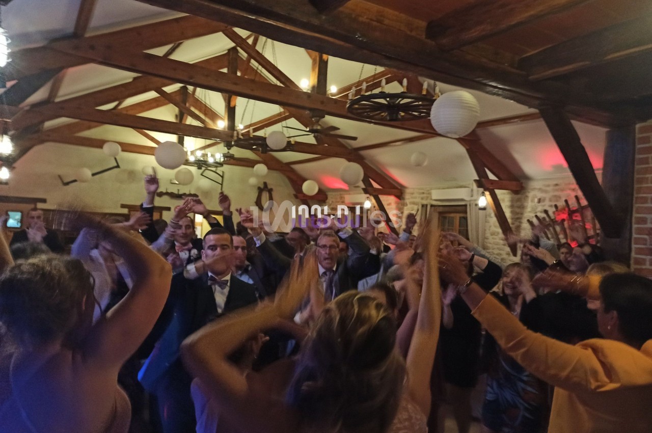 Des personnes dansent et lèvent les bras lors d'une fête dans une salle avec poutres apparentes et décorations suspendues.