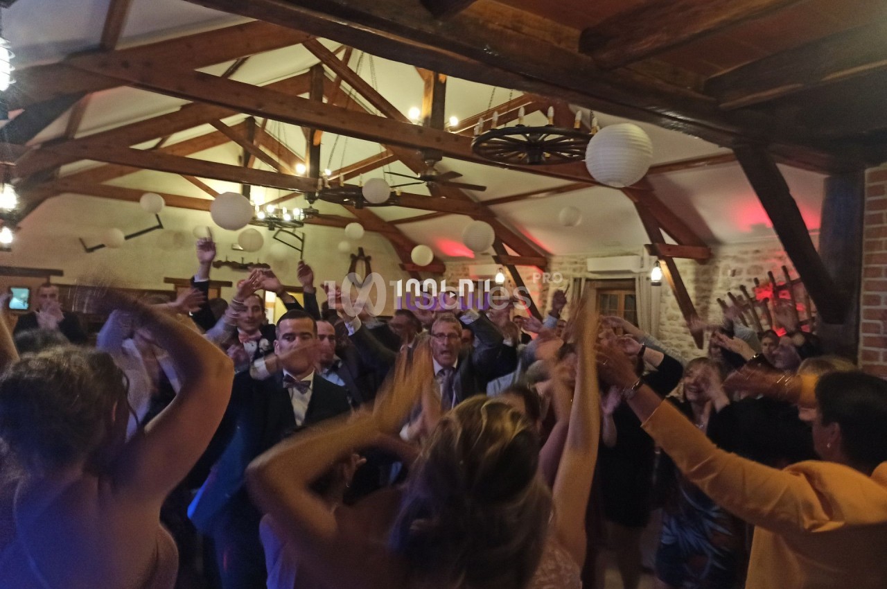 Des personnes dansent et lèvent les bras lors d'une fête dans une salle avec poutres apparentes et décorations suspendues.