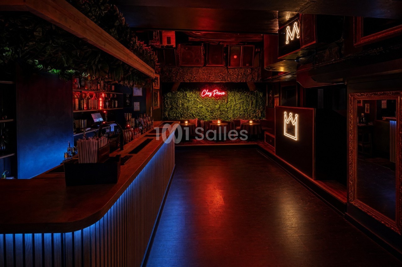 Intérieur d'un bar sombre avec un comptoir éclairé, des néons rouges et une décoration murale végétale.