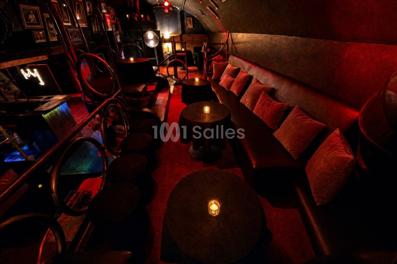 Salle de bar tamisée avec banquettes, coussins, petites tables rondes et éclairage rouge chaleureux.