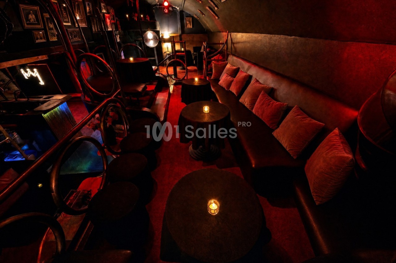 Salle de bar tamisée avec banquettes, coussins, petites tables rondes et éclairage rouge chaleureux.