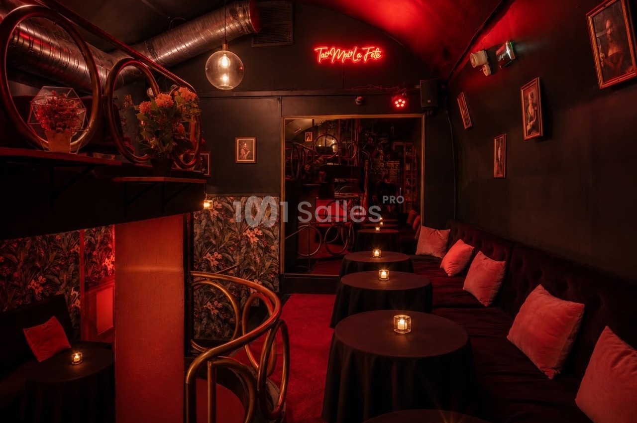 Salle intimiste éclairée par une lumière rouge tamisée, avec tables rondes, bougies et décorations murales.
