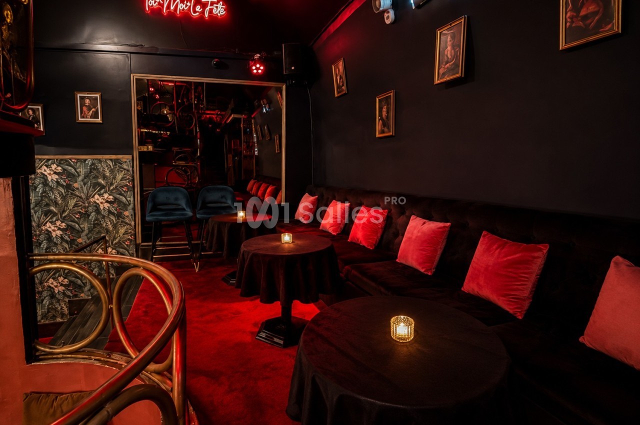 Salle sombre et cosy avec banquettes noires, coussins rouges, tables rondes et bougies, décorée de cadres muraux.