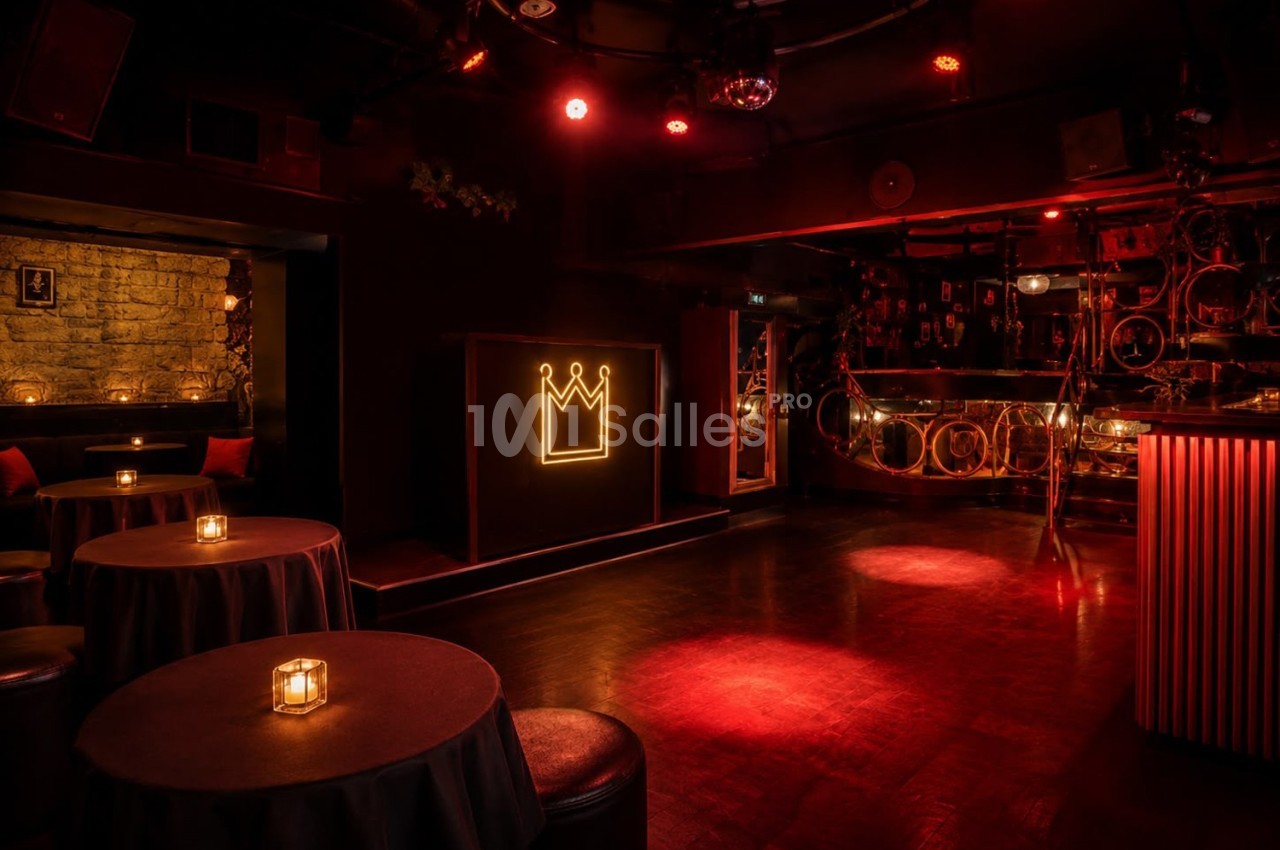 Salle sombre avec éclairage rouge, tables rondes avec bougies, mur en pierre et logo de couronne lumineux.