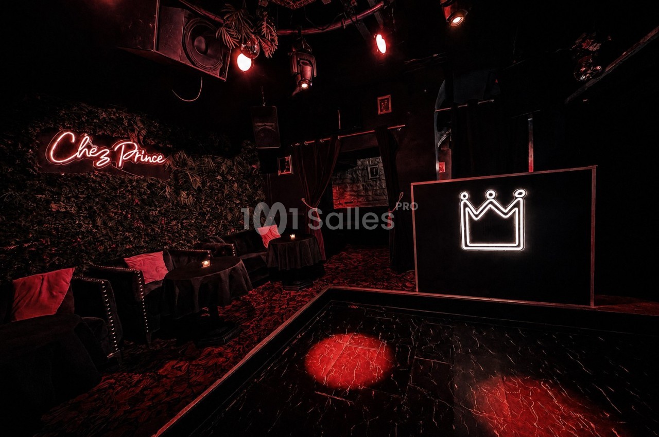 Salle sombre avec éclairage rouge, tables basses, fauteuils, enseigne ’Chez Prince’ et logo de couronne lumineux.