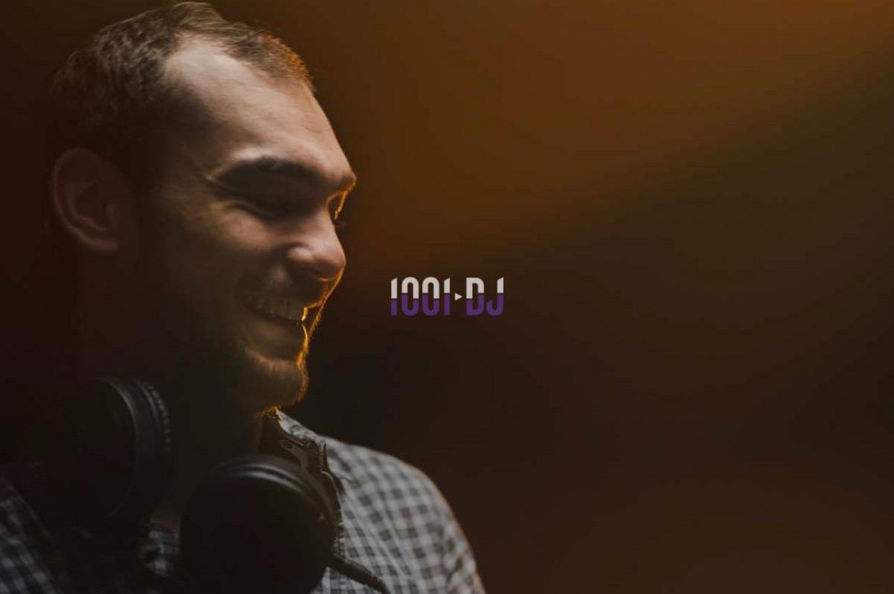 Un homme souriant portant un casque audio autour du cou, éclairé par une lumière chaude sur fond sombre.