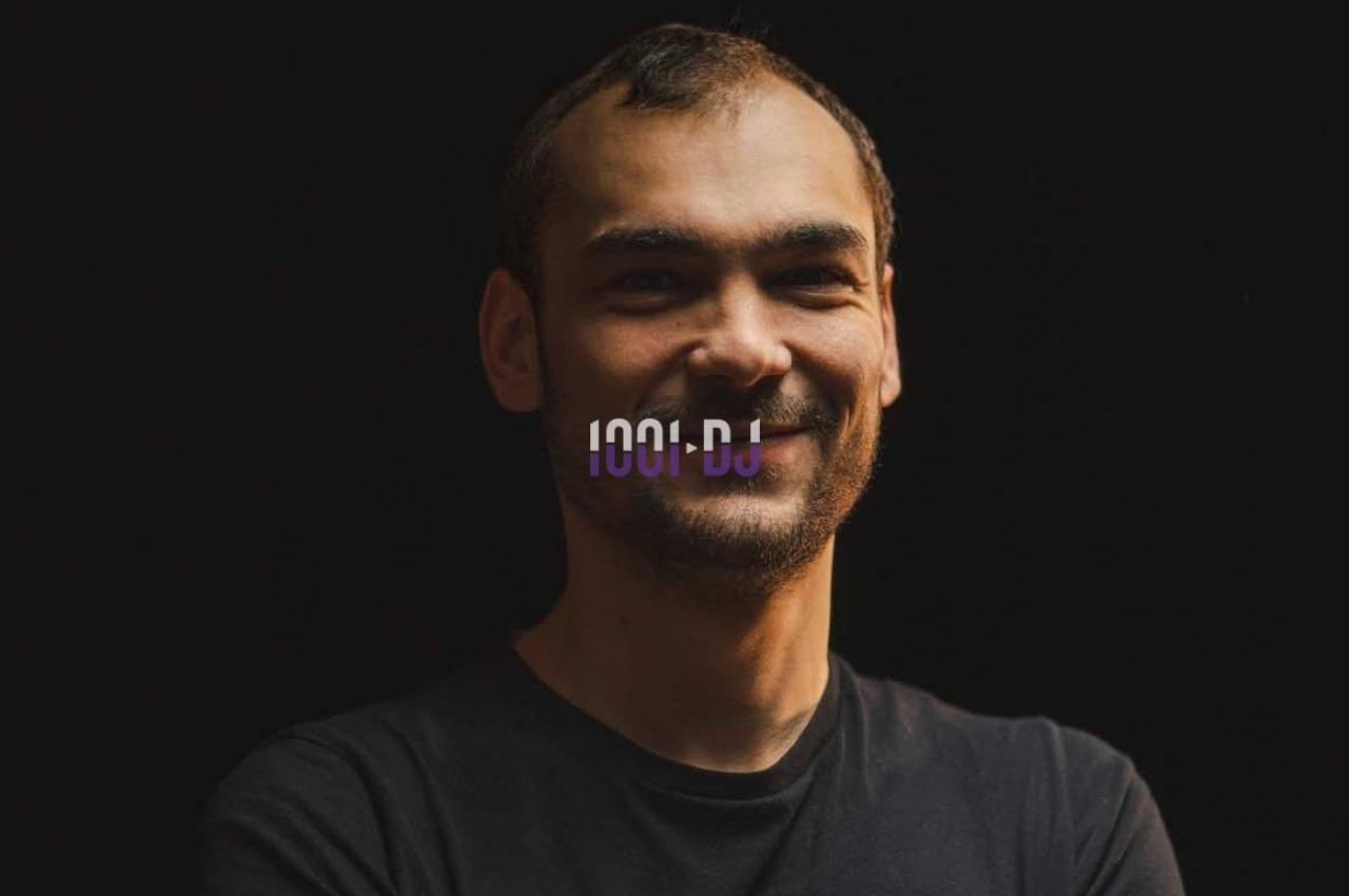 Un homme souriant, portant un t-shirt noir, se tient devant un fond sombre.