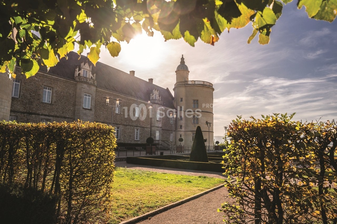 Façade d'un château en pierre entouré de jardins taillés, éclairé par une lumière de fin de journée.