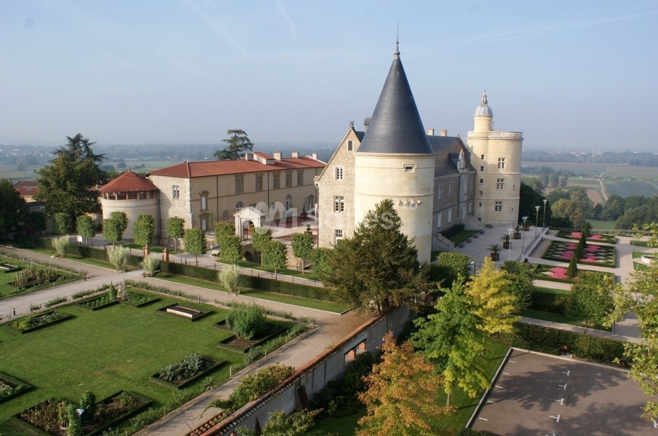 Vue aérienne d'un château entouré de jardins paysagers, avec des allées et des arbres en arrière-plan.