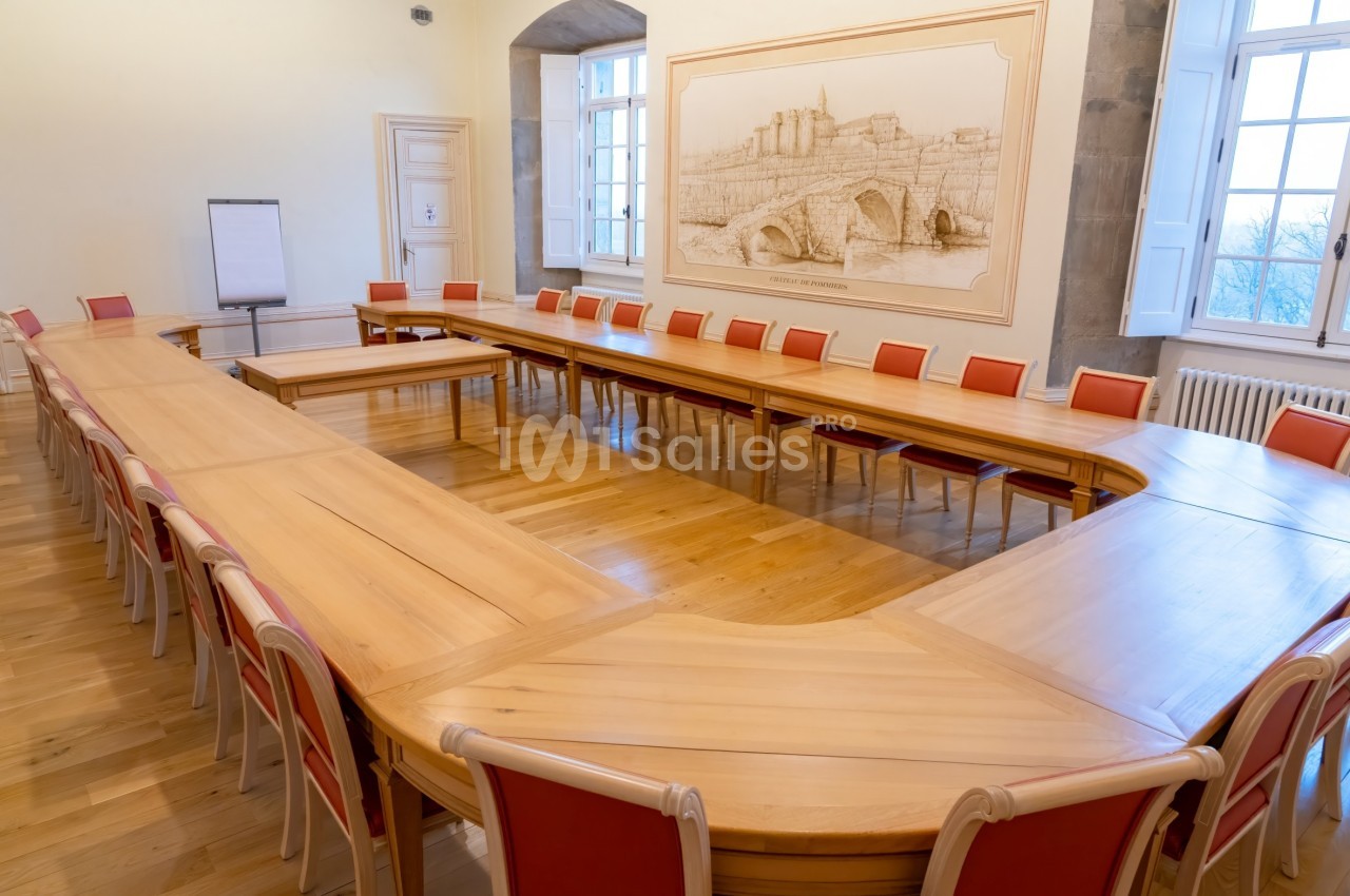 Grande salle de réunion avec une table en U en bois clair, entourée de chaises rouges, et un tableau mural représentant un…