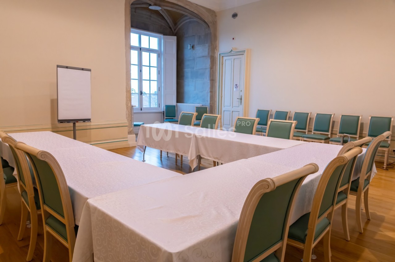Salle de réunion lumineuse avec tables disposées en U, chaises vertes et un tableau blanc sur pied.