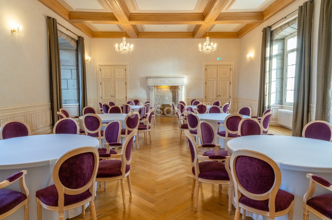 Salle élégante avec parquet, tables rondes couvertes de nappes blanches et chaises violettes, éclairée par des lustres.