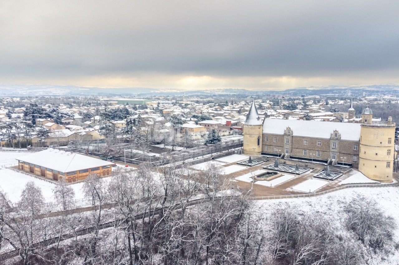 Vue aérienne d'un château entouré de neige, avec une petite ville et un paysage hivernal en arrière-plan.