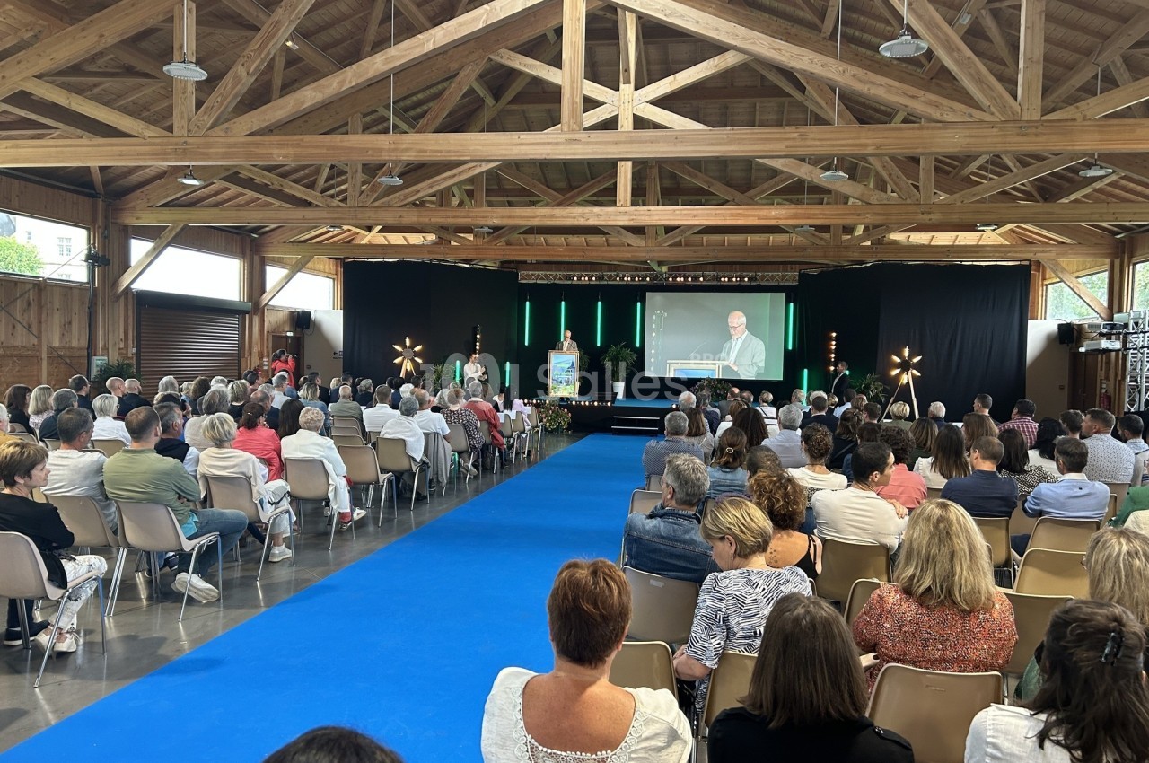 Salle comble avec des participants assis face à une scène où un orateur s'exprime, sous une structure en bois.
