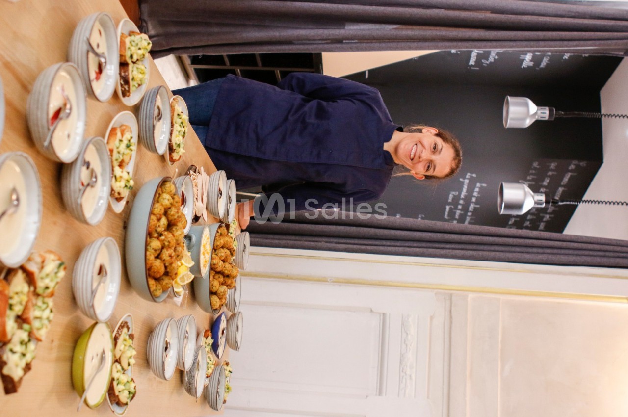 Une personne souriante en tenue de cuisine présente des plats variés disposés sur une table en bois.