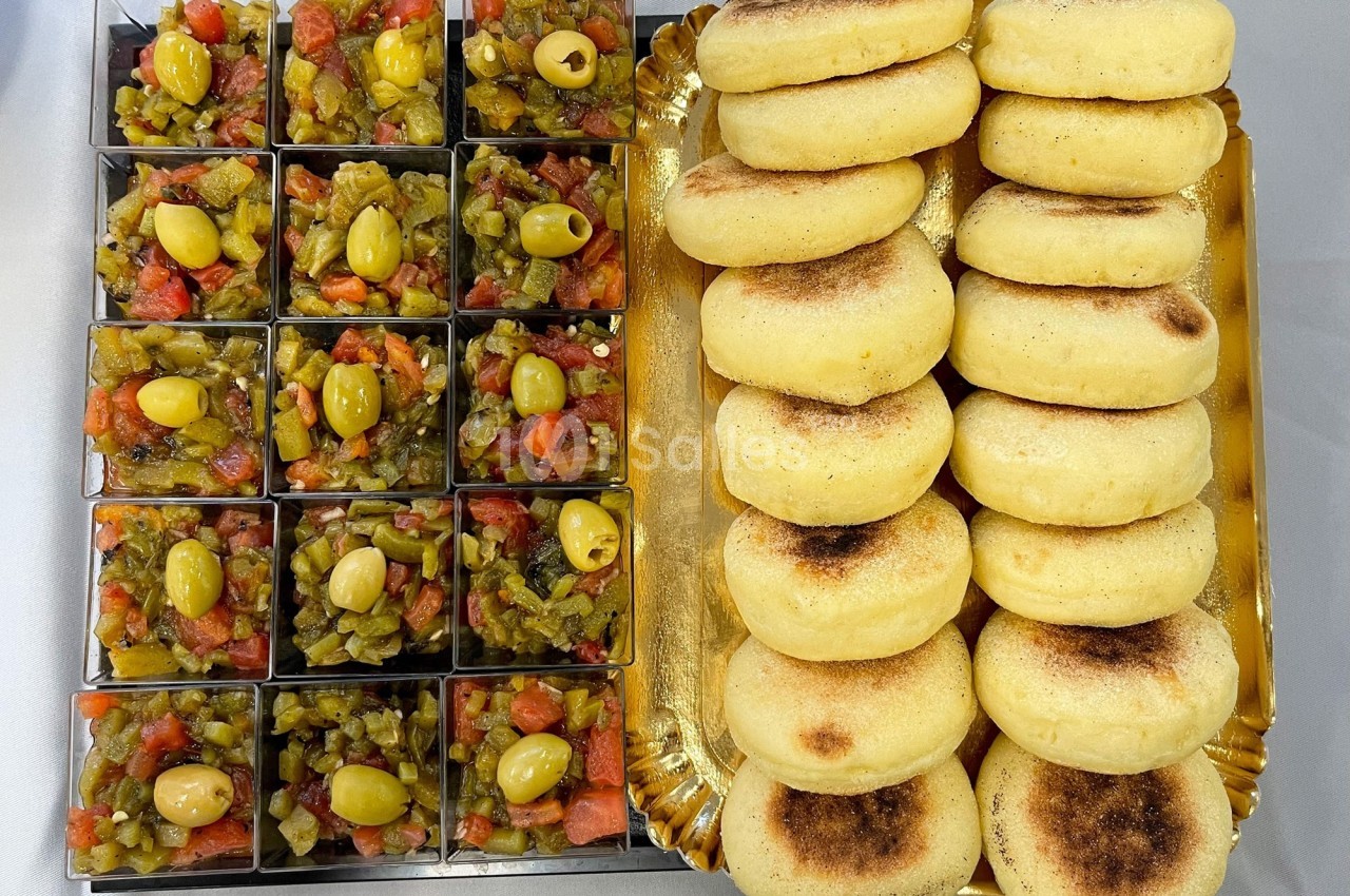 Plateau avec des mini bols de salade de légumes garnis d'olives vertes et des pains ronds dorés empilés.