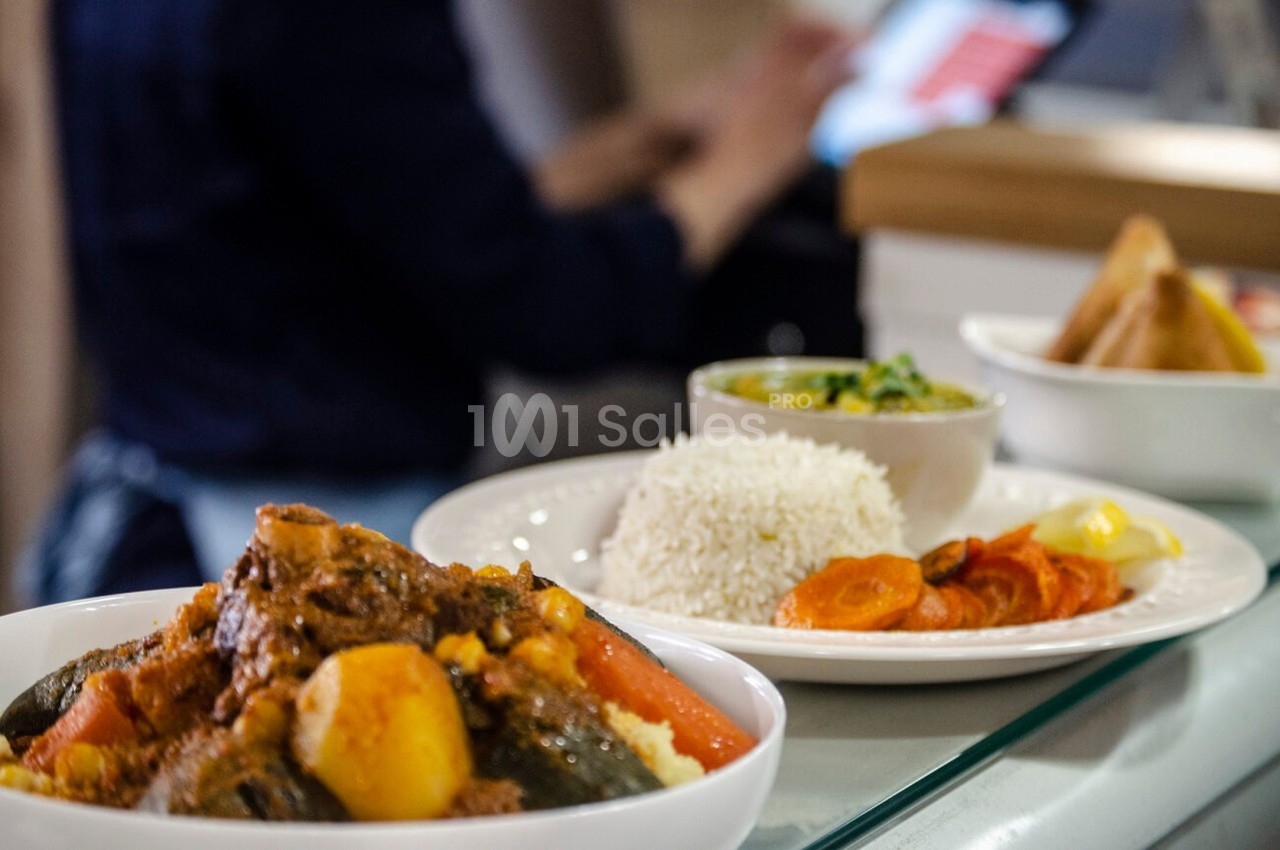 Assiette de couscous avec légumes et viande, accompagnée de riz, carottes et sauce, en arrière-plan une personne floue.