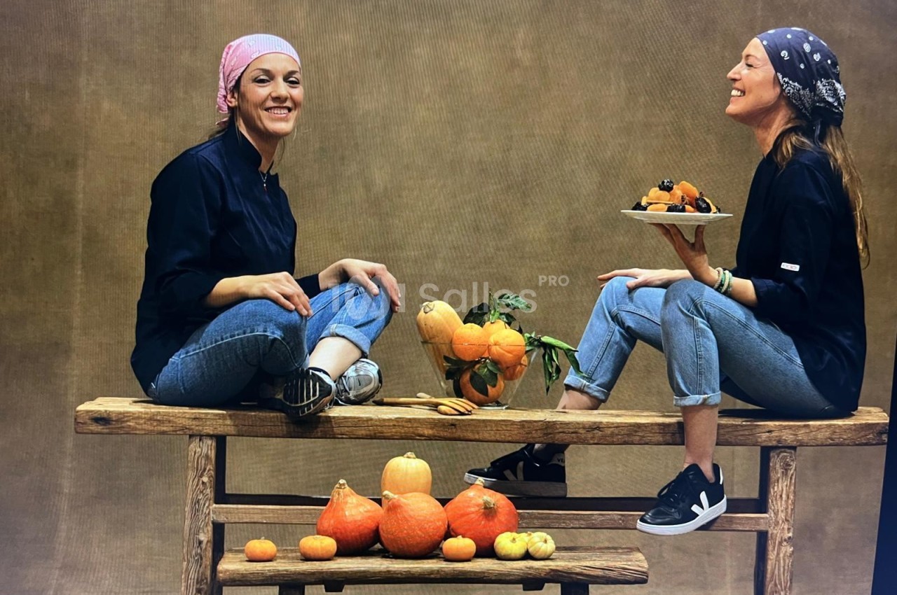Deux femmes assises sur une table en bois, entourées de courges et de fruits, souriant dans un décor neutre.