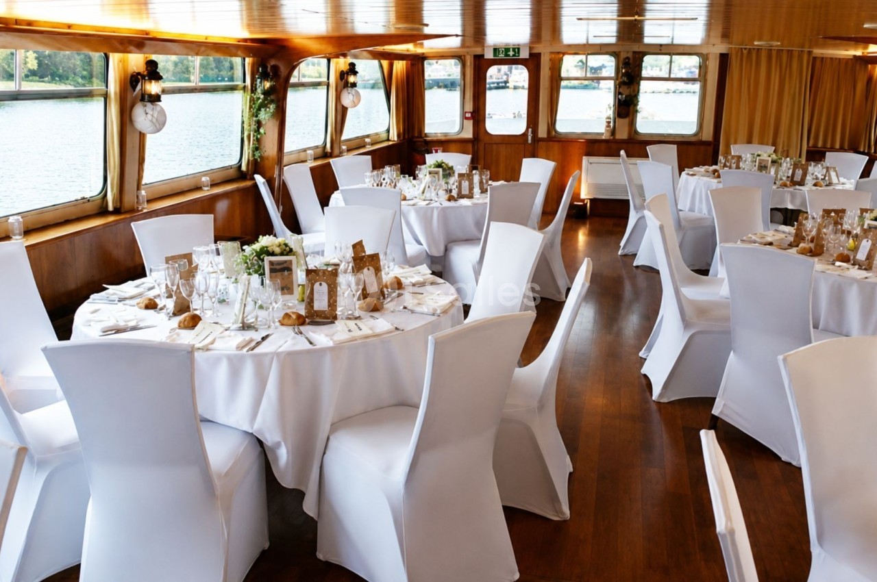 Salle de réception sur un bateau avec des tables rondes dressées, nappes blanches et vue sur l'eau par les fenêtres.