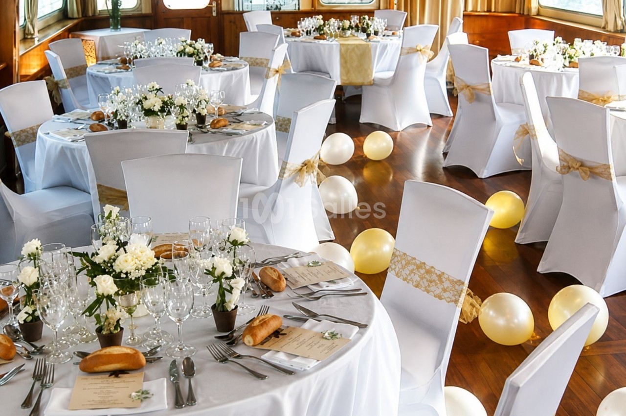 Salle décorée pour un événement avec des tables rondes dressées, nappes blanches, fleurs et ballons dorés au sol.