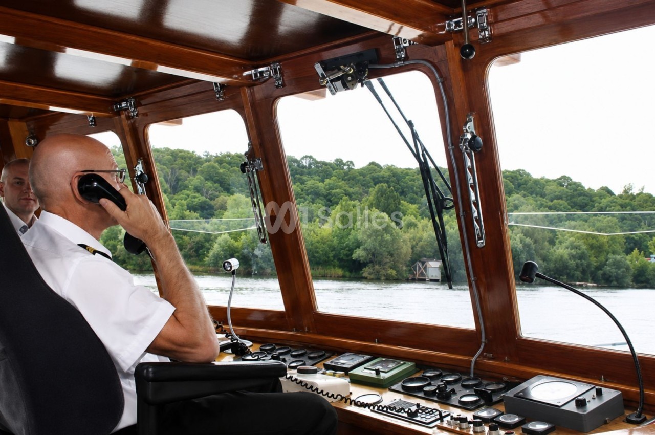 Un capitaine en uniforme utilise une radio dans la cabine d'un bateau, avec vue sur une rivière bordée d'arbres.