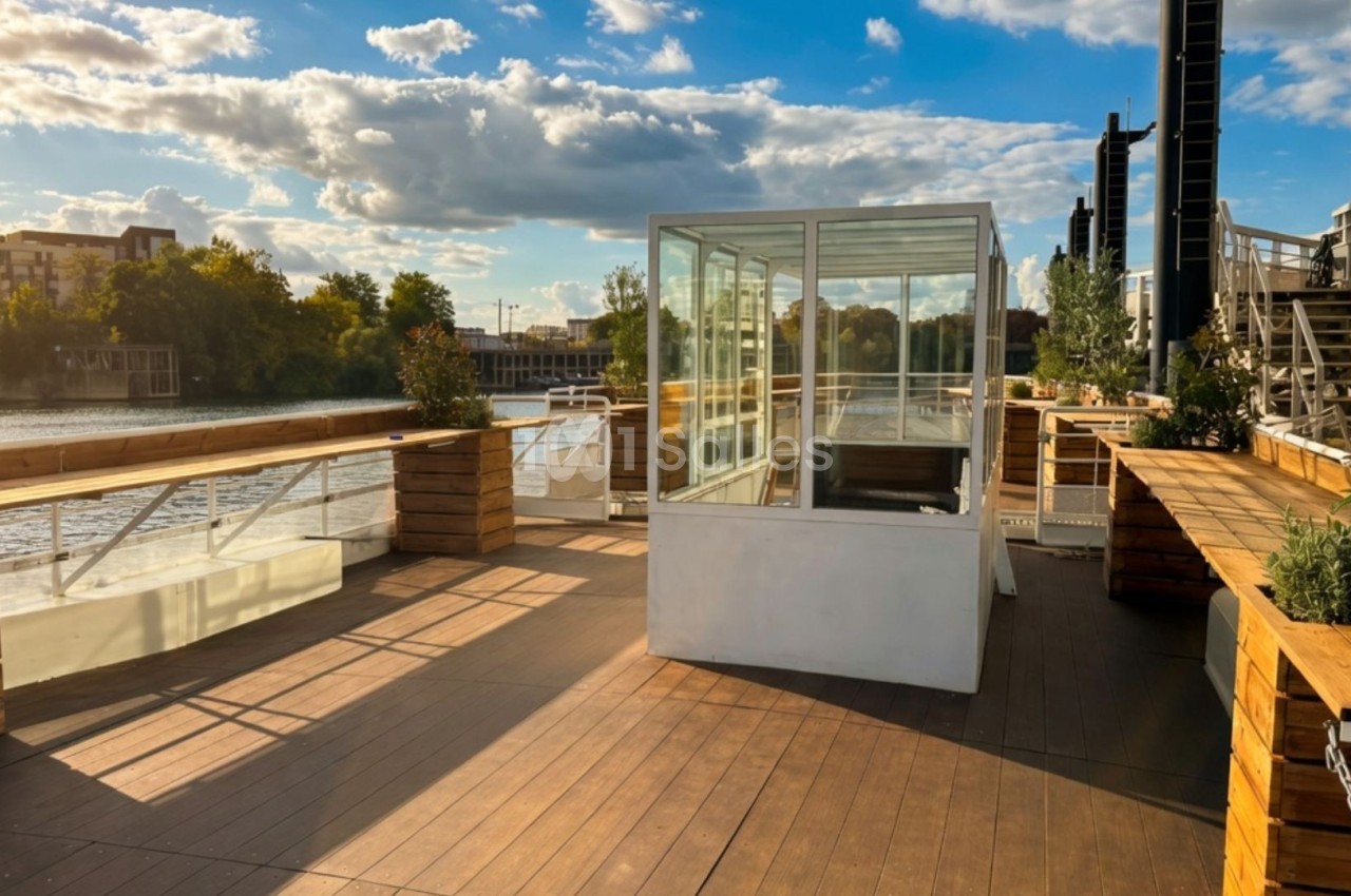 Terrasse en bois avec une petite structure vitrée, bordée de plantes, donnant sur une rivière par temps ensoleillé.