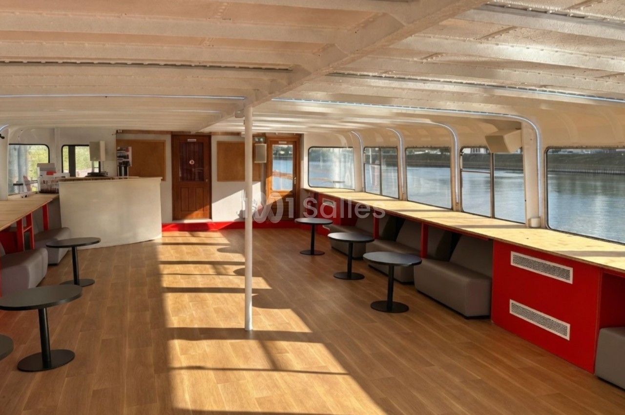 Intérieur lumineux d'un bateau avec des tables, des banquettes rouges et un bar, vue sur l'eau à travers de grandes fenêtres.