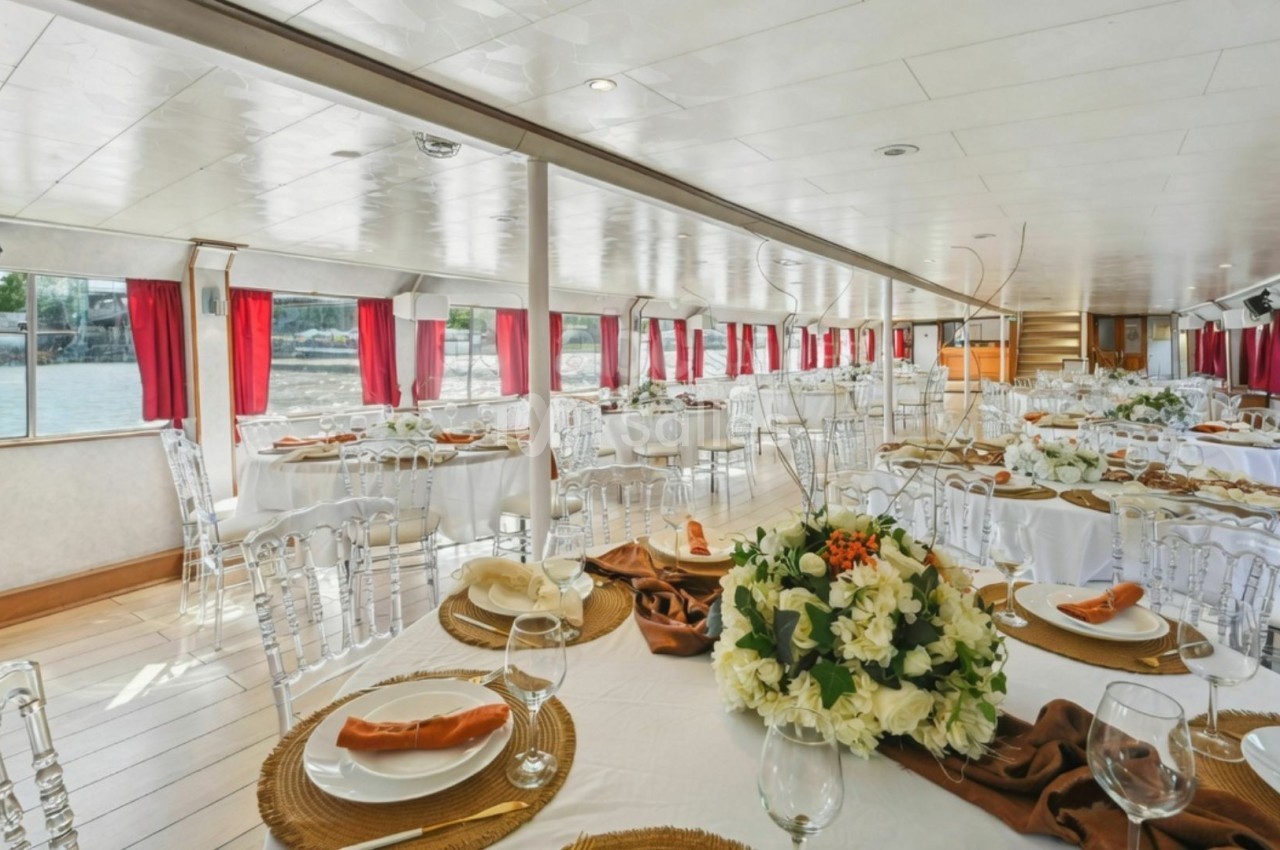 Salle de réception lumineuse sur un bateau, avec tables dressées, nappes blanches et décoration florale élégante.