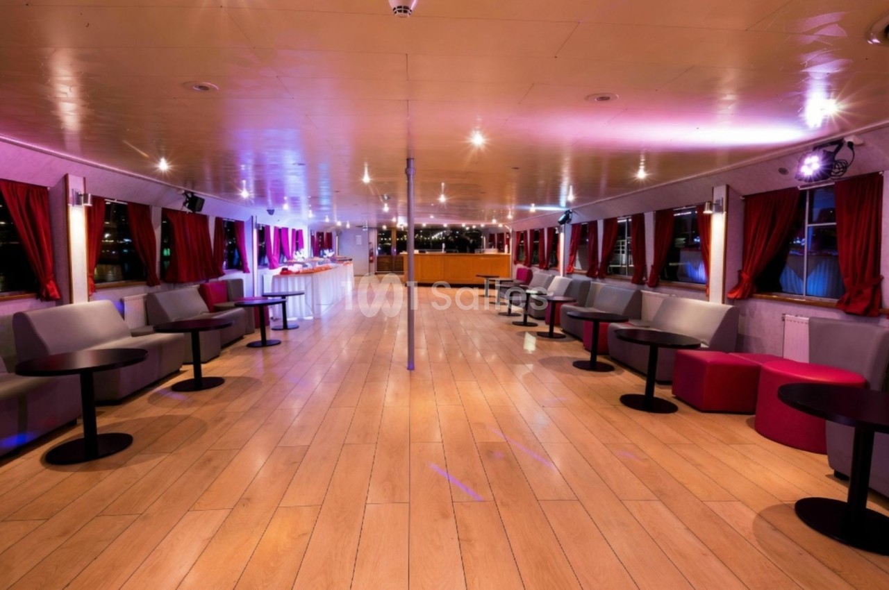 Intérieur d'un bateau avec un sol en bois, des banquettes, des tables basses et des rideaux rouges, éclairage tamisé.