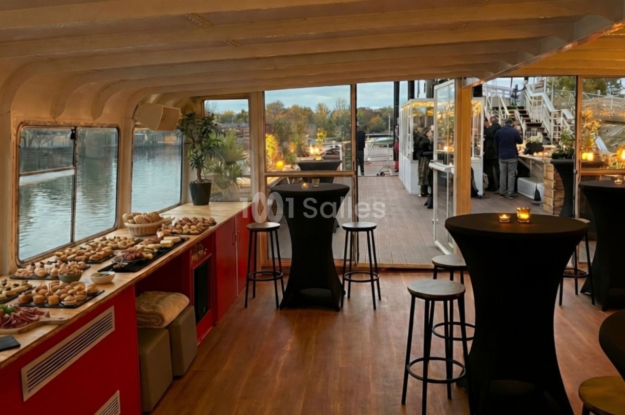Salle intérieure d'un bateau aménagée pour un événement, avec buffet, tables hautes et vue sur une terrasse extérieure.